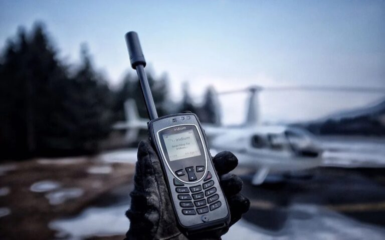 Iridium satellite phone held in a gloved hand beside a small plane in winter